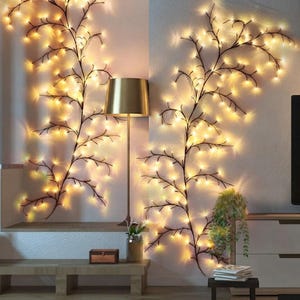 May include: Two decorative wall-mounted light strings shaped like dark, leafless tree branches. Each branch features numerous small, warm-toned LED lights that emit a soft glow. A gold-colored metal floor lamp stands between the two light displays.