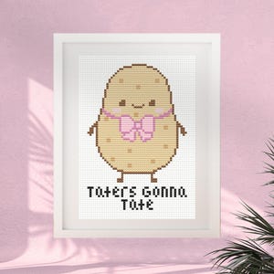 Op de afbeelding: Een ingelijst kruissteekpatroon met een beige aardappelkarakter met een roze strik, kleine armen en benen. De aardappel heeft een lachend gezicht met roze wangen. Onder het figuur staat de tekst Taters Gonna Tate in zwarte letters.