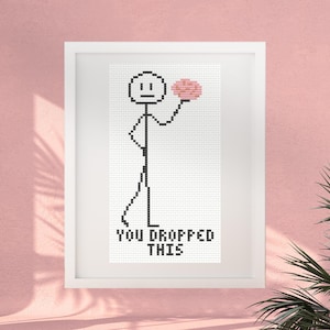 May include: A framed cross-stitch pattern featuring a black stick figure on a white background. The figure holds a pink brain-shaped object in one hand. Below the figure, the text reads YOU DROPPED THIS in black, blocky, pixelated letters.