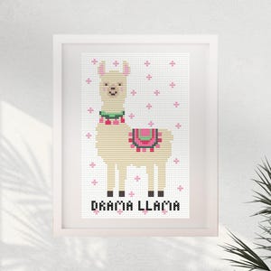 May include: A framed cross-stitch pattern featuring a beige llama with a green collar and a pink and red saddle. The background is white with small pink cross-stitch accents. The text DRAMA LLAMA is printed in black at the bottom of the design.
