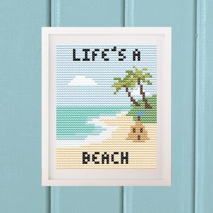 May include: A framed cross-stitch artwork featuring a beach scene with two green palm trees, a tan sandcastle with a blue flag, and the black text "LIFE'S A BEACH" against a light blue sky and turquoise ocean background in a white frame.