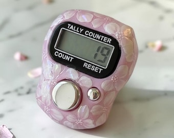 Könnte beinhalten: Ein hellrosa digitaler Zähler mit weißem Blumenmuster und irisierender Oberfläche. Das Gerät verfügt über ein schwarzes LCD-Display, das die Zahl 19 anzeigt, sowie die in Weiß auf den schwarzen Rahmen gedruckten Begriffe TALLY COUNTER, COUNT und RESET.
