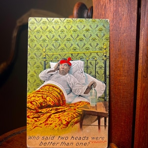 May include: A vintage postcard featuring a person in bed with a red bandage on their head, holding a glass of water. The illustration includes a green patterned wallpaper, a metal bed frame, and the text: Who said two heads were better than one?