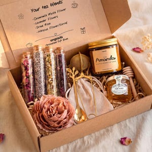 Könnte beinhalten: Eine Geschenkbox mit vier Glasfläschchen gefüllt mit getrockneten Blumen und Kräutern, einem goldenen Tee-Ei, einem goldenen Löffel, einem Glas mit der Aufschrift For Amanda, einem kleinen Honigglas, einem Honiglöffel aus Holz und einer großen künstlichen rosa Blume.