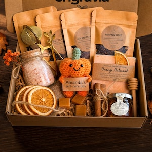 May include: A gift box containing three tea pouches, a gold tea infuser, a glass jar of pink bath salts, a crocheted orange pumpkin holding a sign that reads Amanda's self-care day, orange oatmeal soap, dried orange slices, caramels, cinnamon sticks, and honey.