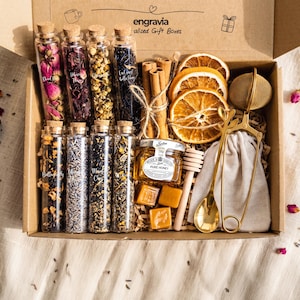 May include: A gift box containing eight glass vials filled with dried flowers and tea leaves, three dried orange slices, a bundle of cinnamon sticks, a small jar of honey, three honey candies, a wooden honey dipper, and a gold tea infuser spoon.