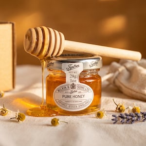 May include: A small glass jar of Tiptree Pure Honey with a silver lid and a white label reading Wilkin & Sons Ltd, Tiptree, Essex, England, and Net Wt. 28g 1oz. A wooden honey dipper rests on top, with golden honey dripping down the side of the jar.