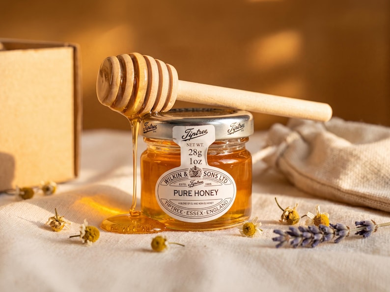 May include: A small glass jar of Tiptree Pure Honey with a silver lid and a white label reading Wilkin & Sons Ltd, Tiptree, Essex, England, and Net Wt. 28g 1oz. A wooden honey dipper rests on top, with golden honey dripping down the side of the jar.
