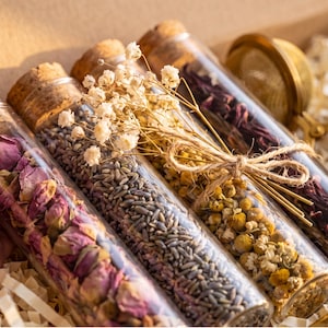 May include: Four clear glass test tubes with cork stoppers, each filled with different dried botanicals including pink rose buds, lavender, chamomile flowers, and dark red hibiscus petals, tied together with twine and small white flowers.
