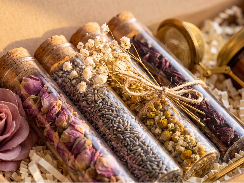 May include: Four clear glass test tubes with cork stoppers, each filled with different dried botanicals including pink rose buds, lavender, chamomile flowers, and dark red hibiscus petals, tied together with twine and small white flowers.