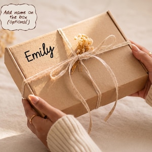 May include: A rectangular brown cardboard gift box featuring the name Emily printed in black script. The box is tied with thin twine and decorated with a small sprig of dried flowers. A text bubble above the box reads Add name on the box (optional).