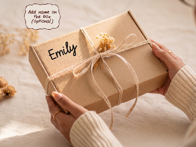 May include: A rectangular brown cardboard gift box featuring the name Emily printed in black script. The box is tied with thin twine and decorated with a small sprig of dried flowers. A text bubble above the box reads Add name on the box (optional).