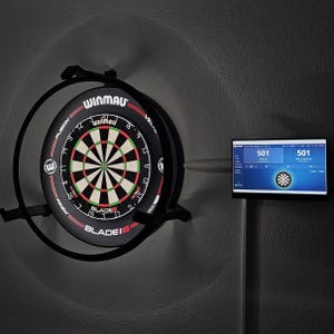 May include: A Winmau Blade 6 dartboard mounted within a black circular frame, illuminated by a ring light. Beside it, a digital display screen shows a dart scoring interface with the number 501 visible on a blue background, mounted on a stand.