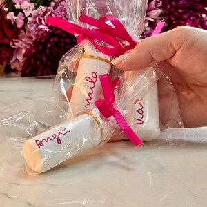 May include: Three white cylindrical lip balm tubes individually wrapped in clear plastic bags, each tied with a bright pink ribbon. The tubes feature gold-colored bands and are personalized with the names Angie, Mila, and Kat in pink script.