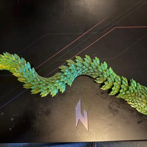 May include: A flexible, articulated toy snake featuring a vibrant green and yellow scale-like texture throughout its entire body. The segmented design allows the creature to curve and bend into various positions, showcasing a detailed, layered appearance.