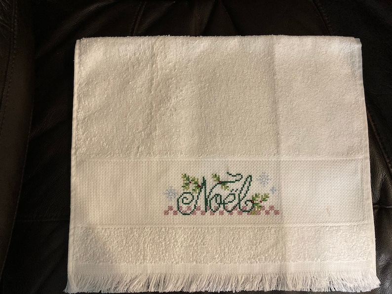 May include: White hand towel with a green and red embroidered design that says "Noel" with snowflakes and holly.
