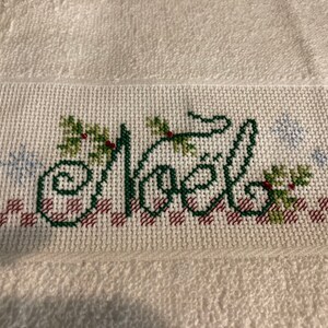 May include: White towel with a green embroidered word "Noel" with red berries and green leaves. The word is surrounded by a red and white checkered pattern and snowflakes.