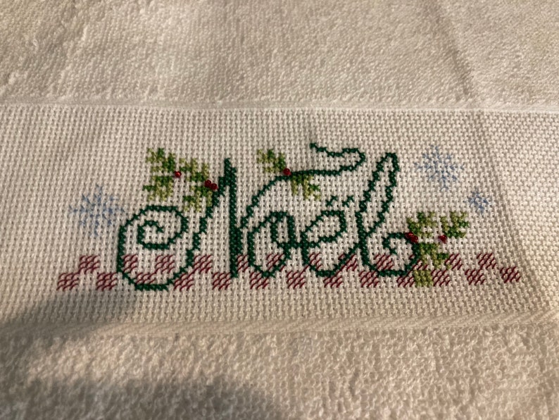 May include: White towel with a green embroidered word "Noel" with red berries and green leaves. The word is surrounded by a red and white checkered pattern and snowflakes.
