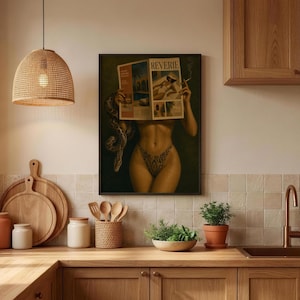 Op de afbeelding: Een ingelijste kunstdruk van een persoon die een tijdschrift genaamd REVERIE vasthoudt, met een slang om de arm gewikkeld. De persoon draagt een bikinibroekje met patroon. Het kunstwerk hangt in een keuken met houten kasten en een rieten lamp.