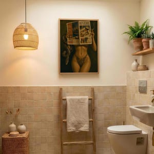 Op de afbeelding: Een ingelijste kunstdruk met de titel REVERIE met een figuur die een tijdschrift vasthoudt, een hanglamp van gevlochten rotan, een houten handdoekladder met een beige handdoek, een rieten opbergmand en varens op een houten plank in een badkamer.
