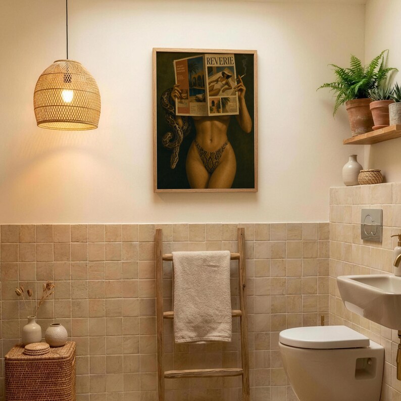 Op de afbeelding: Een ingelijste kunstdruk met de titel REVERIE met een figuur die een tijdschrift vasthoudt, een hanglamp van gevlochten rotan, een houten handdoekladder met een beige handdoek, een rieten opbergmand en varens op een houten plank in een badkamer.