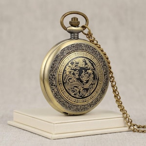 May include: A round antique brass pocket watch featuring a black circular center inlay with a gold dragon illustration. The watch case has an intricate engraved border pattern and includes a matching brass chain attached to the top winding crown.