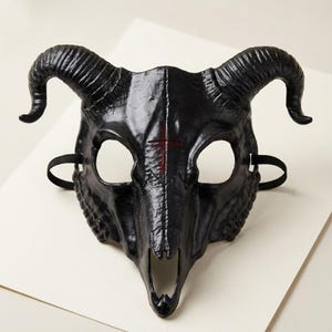 May include: A black ram skull mask featuring large curved horns, two eye openings, and a nasal cavity opening. A thin red cross is painted vertically between the eye sockets. The mask includes black elastic straps on the sides for securing it.