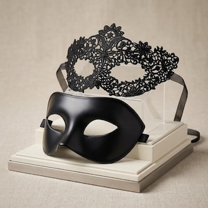 May include: A set of two masquerade masks. One mask is crafted from intricate black lace with a floral pattern and features black ribbon ties. The second mask is a smooth, solid matte black mask with a simple, curved design and side straps.