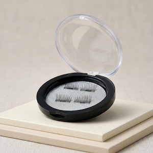May include: A round black plastic compact case with a clear hinged lid, containing two pairs of black false eyelashes resting on a textured silver circular base. The compact is open, displaying the lashes clearly against the metallic background.
