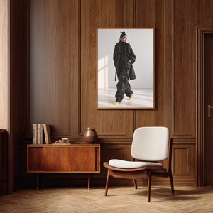 Op de afbeelding: Een ingelijste wandkunstprint met een persoon in een zwart jack met lange mouwen en een bijpassende zwarte broek met felgele puntige schoenen, tentoongesteld in een kamer met donkerhouten lambrisering en een witte gestoffeerde stoel.