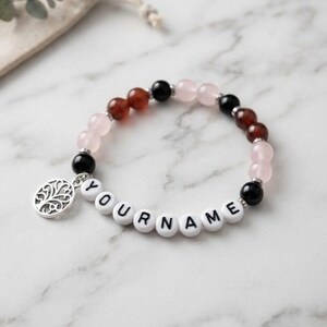 May include: A beaded bracelet featuring alternating round black, translucent pink, and reddish-brown beads. The bracelet includes white spherical beads with the black printed text YOURNAME and a silver-toned circular charm with a tree design.