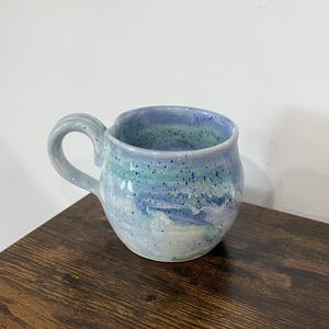 May include: A round ceramic mug featuring a marbled glaze in shades of light blue, lavender, and white. The surface is decorated with small dark blue and yellow speckles throughout. It has a curved handle and a smooth, glossy finish.