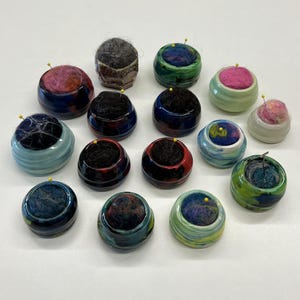 May include: A collection of fourteen small, round ceramic pin cushions with glazed, multi-colored surfaces. Each container holds a soft, felted wool insert in various shades of black, pink, blue, and green, topped with a single yellow pin.
