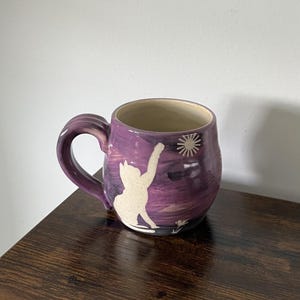 May include: A purple ceramic mug featuring a white silhouette of a cat reaching toward a sun or star graphic. The mug has a rounded body, a thick handle, and a cream-colored interior. The exterior surface displays a hand-painted, streaked finish.