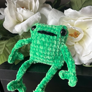 May include: A small, bright green plush frog made of soft, textured yarn. The frog has a square-shaped body, long thin arms and legs, and two large, round, black plastic eyes. It features a simple, straight black stitched mouth.