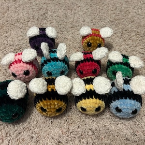 May include: A collection of ten small, round, crocheted bee plushies made from soft, fuzzy yarn. Each bee features black stripes, two white crocheted wings, and two black plastic safety eyes. The bees come in various colors including yellow, pink, purple, teal, red, green, dark green, and light blue.