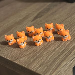 May include: A collection of eleven small, orange and white fox figurines. Each miniature fox features pointed ears, a white chest, and black painted eyes and nose. The figures are arranged in a group, each depicted in a seated position.