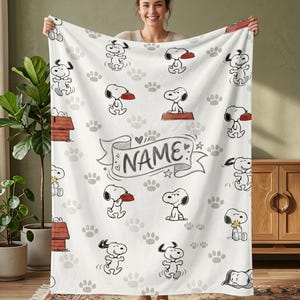 May include: A white fleece blanket featuring a repeating pattern of Snoopy in various poses, including sitting on a red doghouse and holding a red food bowl. The centre displays a banner with the word NAME in black text, surrounded by grey paw prints.