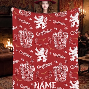 Puede incluir: Una manta de forro polar roja con un patrón blanco repetitivo del escudo del león de Gryffindor, la palabra Gryffindor e iconos de snitch dorada. La manta incluye una zona de texto blanca en la parte inferior con la palabra NAME en fuente estilizada.