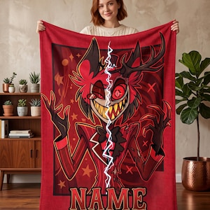 May include: A rectangular red fleece blanket featuring a graphic illustration of a character with antlers, a wide grin, and a red suit. A vertical white lightning bolt divides the character, and the word NAME is printed in bold letters at the bottom.