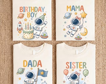 Puede incluir: Cuatro camisetas de color blanco roto con gráficos de temática espacial. La primera dice BIRTHDAY BOY con un astronauta y un pastel. La segunda MAMA con un astronauta y un globo de estrella. La tercera DADA con un astronauta y una bandera. La cuarta SISTER con un astronauta en un cohete.