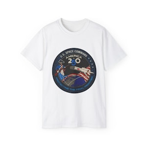 May include: A white short-sleeved t-shirt featuring a circular graphic with a colonial soldier, a rocket, and the text U.S. SPACE COMMAND AMERICA 250 PROTECTING THE FINAL FRONTIER. The design includes red, white, and blue colours on a dark background.