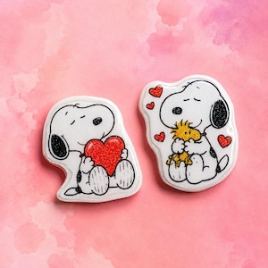 Pode incluir: Dois pins brancos do personagem Snoopy com contornos pretos. O pin da esquerda mostra o Snoopy segurando um grande coração vermelho com glitter. O pin da direita mostra o Snoopy abraçando um personagem amarelo de Woodstock com glitter.