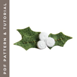 May include: A felt holly sprig with three white, spiral-shaped berries. The green leaves have embroidered details and a gold-colored stem. The text "PDF PATTERN & TUTORIAL" is on the left side of the image.