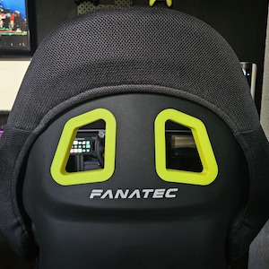 May include: A black racing-style gaming chair featuring a mesh headrest, two lime green harness pass-through cutouts, and the white Fanatec logo printed on the center of the backrest. The chair has a contoured frame and a textured fabric finish.