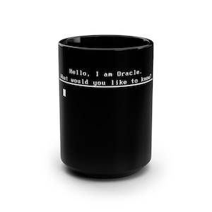 Può includere: Una tazza in ceramica nera con testo bianco in stile terminale informatico che recita: Hello, I am Oracle. What would you like to know? Il design include una linea orizzontale e un piccolo cursore quadrato bianco posizionato sotto il testo.