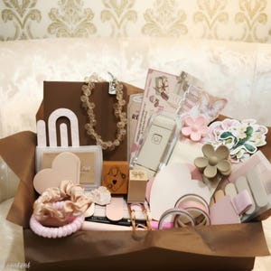 May include: A gift box filled with stationery and accessories including a beige beaded necklace, a small portable label maker, heart-shaped paper cutouts, a flower-shaped hair clip, pink scrunchies, washi tape rolls, and various stickers.