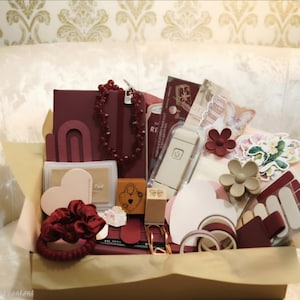 Op de afbeelding: Een geschenkdoos gevuld met briefpapier en accessoires in bordeauxrood, crème en beige tinten. De items bevatten een donkerrood notitieboek, een kralenketting, een wit elektronisch apparaat, hartvormige kaarten, bloemenstickers en een haarelastiek.