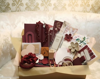 May include: A gift box filled with stationery and accessories in shades of burgundy, cream, and beige. Items include a dark red notebook, a beaded necklace, a white electronic device, heart-shaped cards, floral stickers, a hair scrunchie, and tape.