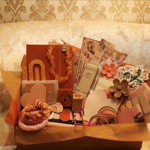 May include: A gift box filled with stationery and accessories in warm earth tones, including a notebook, a beaded necklace, a portable label maker, heart-shaped cards, floral stickers, hair clips, a scrunchie, and various rolls of adhesive tape.